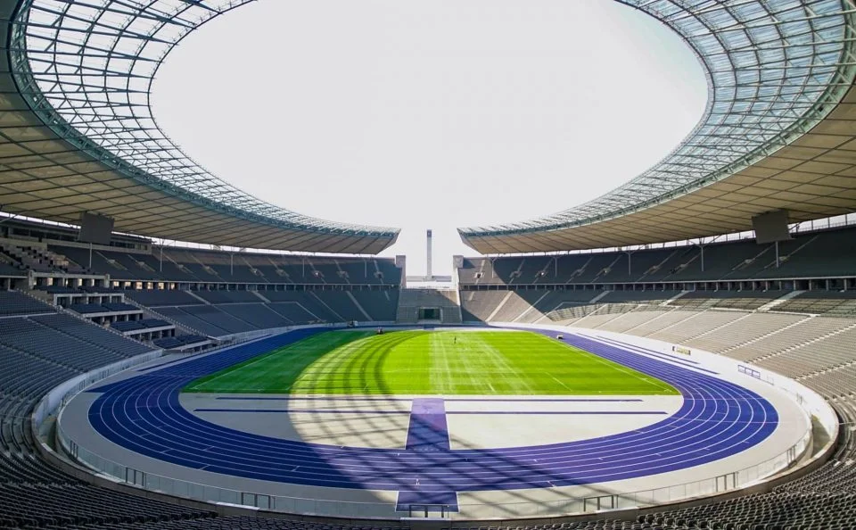 An empty track and field area with a blue track surrounding a green field, enclosed roof, and stadium seating.