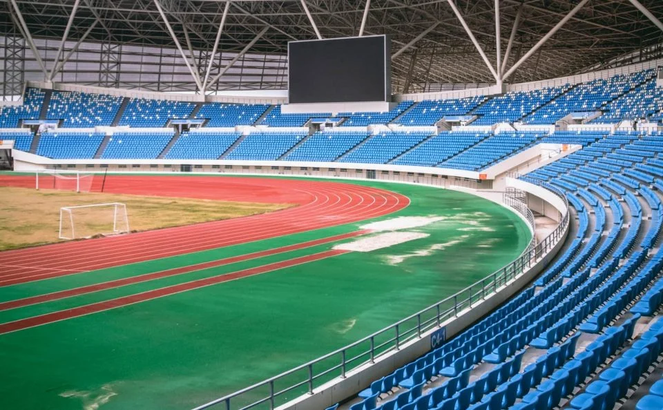An empty track and field area with a red track surrounding a green field, enclosed roof, and blue stadium seating.