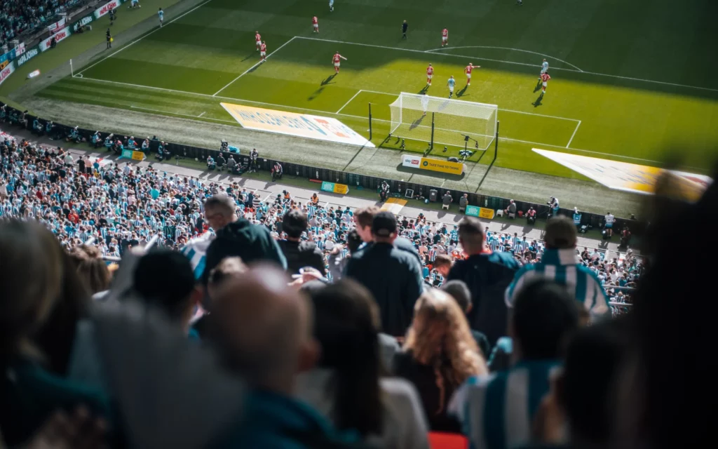 Fans watch a soccer game from bleacher seating in a packed stadium.